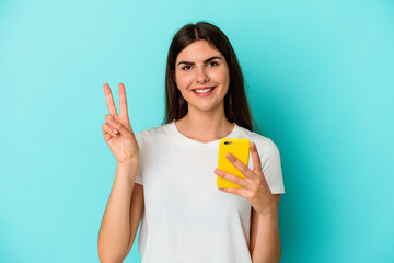 Young caucasian woman holding a mobile phone isolated on blue background joyful and carefree showing a peace symbol with fingers.