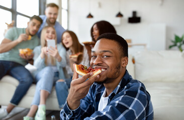 Handsome black guy eating slice of pizza, group of his multiethnic friends with smartphone on background