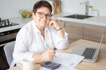 Serious mature Caucasian 60s woman sit at table calculate household finances paying bills on laptop...