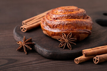 Cinnamon roll on a brown cutting board with anise star and cinnamon sticks around it. Wooden table, close up.