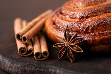 Close up view of crispy cinnamon bun, anise star, cinnamon sticks on brown cutting board. Bakery, sweet dessert, aromatic food concept.