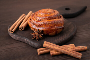 Brown cutting board on dark wooden table, sweet bun, cinnamon sticks and anise star on it. Rustic...