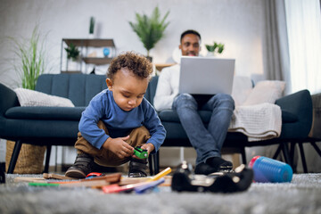 Cute african boy playing with toys on soft carpet while his father working on portable computer on background. Young man taking care of son and working remotely from home.
