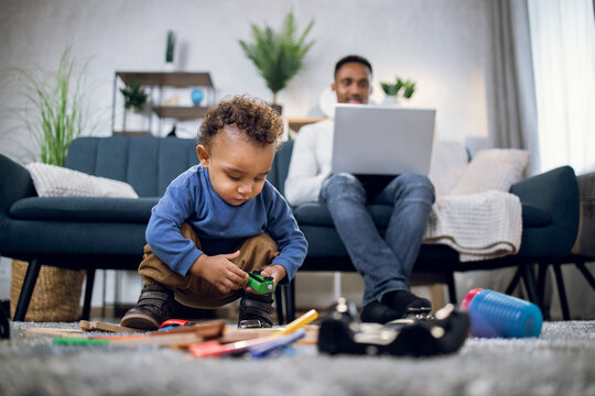 Cute African Boy Playing With Toys On Soft Carpet While His Father Working On Portable Computer On Background. Young Man Taking Care Of Son And Working Remotely From Home.