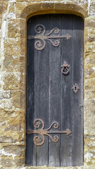 Detail of a medieval weathered oak plank door, with intricate lattice ironwork door fittings. Set into a champhered return local stone wall. Portrait image with texture. Oxfordshire, England. - 431788863