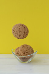 Flying brown round cookies and some cookies lie in little transparent plate on  white wooden table and bright yellow background, creative food concept
