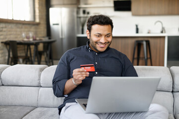 Happy indian guy holding credit card and paying online, using laptop computer for shopping online,...