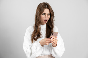 Naklejka premium Surprised, pretty girl is very surprised, looking at a mobile phone. The charming girl in glasses is shocked by what she saw in her phone.