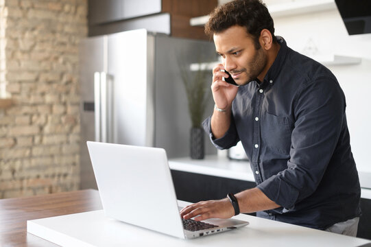 Busy mixed-race young man in smart casual shirt using laptop computer for remote work, a guy is talking on the phone and typing in same time standing in the kitchen at home, work on the distance