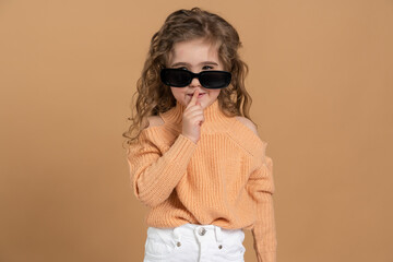 Attractive little girl in sunglasses shows a sign of silence. Girl touches the finger of her lips on a brown background