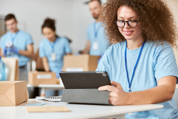 Young woman, volunteer in blue uniform using tablet pc and smiling while sitting indoors. Team sorting, packing items in the background