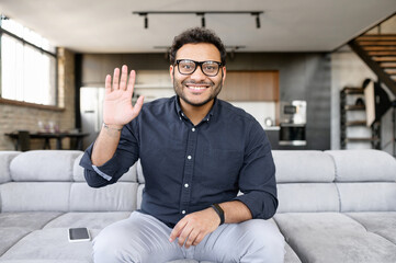 Video chat with indian male employee, mixed race colleague. Screen with a mixed-race guy on it, a man in eyeglasses and shirt sits on the couch looks at camera and waving, video meeting, video call