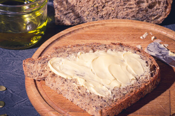 Slice of rustic natural yeast-free bread with flax, poppy seeds, sesame seeds, millet, pumpkin and sunflower seeds, spread with butter, on a wooden board, with olive oil in a glass jar, black