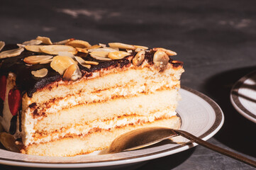 Piece of layered homemade sponge cake on a white saucer with a spoon and a linen napkin, black background, hard light, photo in a low key.