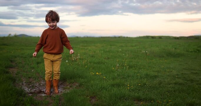 happy redhead boy, kid having fun on a spring meadow, jumping in muddy puddle