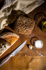 Still life - yeast-free buckwheat bread, black bread with sunflower seeds, olive oil and coarse salt in glass jars, a knife, and a linen napkin on a wooden board, wooden background, hard light, photo