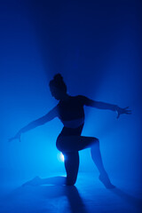 Fototapeta na wymiar Professional ballerina dancing ballet in smoke. Female in black bodysuit on floodlights background.