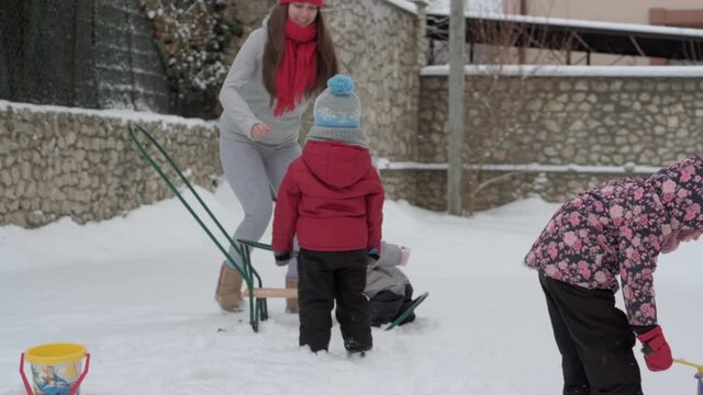 vacation, game, family concept - slo-mo authentic three happy preschool toddler kids siblings with young wonan mom sledding and play with snow. snowfall in cold season weather in winter yard outdoors