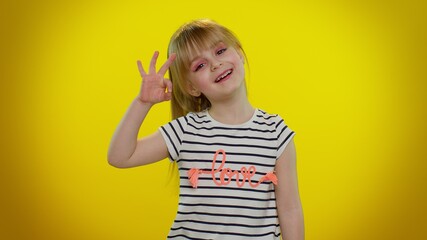 Little blonde teen child kid girl in t-shirt looking approvingly at camera showing ok gesture, like sign positive something good, isolated on yellow studio background. Childhood. Young children
