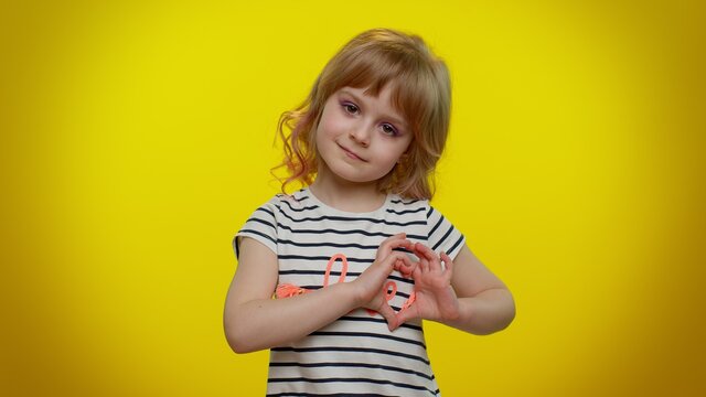 Portrait of smiling blonde child girl 5-6 years old in striped t-shirt makes heart gesture demonstrates love sign expresses good feelings and sympathy on yellow studio background. Teen kid children