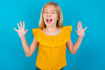 Emotive Caucasian kid girl wearing yellow T-shirt against blue wall laughs loudly, hears funny joke...