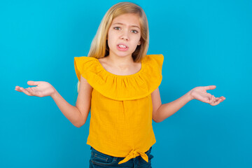 Clueless Caucasian kid girl wearing yellow T-shirt against blue wall shrugs shoulders with...