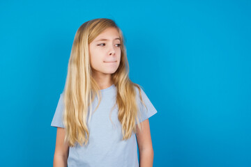 Amazed puzzled beautiful Caucasian little girl wearing white T-shirt over blue background, curves lips and has worried look, sees something awful in front.