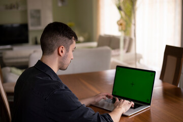 Junger Mann mit schwarzen Haaren sitzt am Tisch in Wohnzimmer und tippt auf dem Laptop. Display ist grün, green screen. Die Möbel ist weiß, grüne Wände. Er ist Student oder Schuler, Home Office, Shule