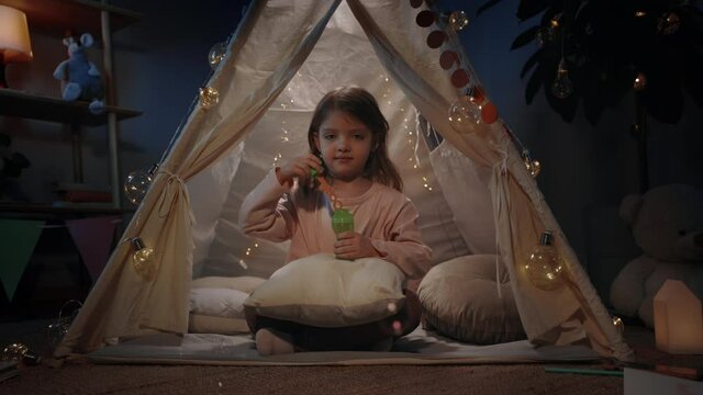 Pretty girl in beautiful makeshift blowing bubbless and looking to camera at home in evening. Little kid sitting on floor while having fun. Concept of leisure and careless childhood.