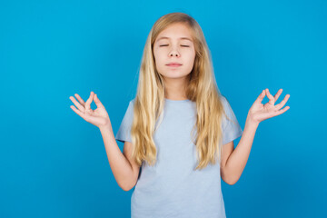 Fototapeta premium beautiful Caucasian little girl wearing blue T-shirt over blue background doing yoga, keeping eyes closed, holding fingers in mudra gesture. Meditation, religion and spiritual practices.