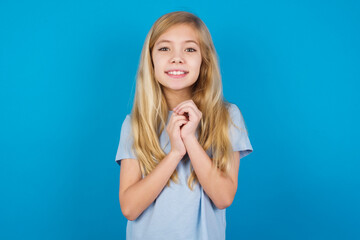 Dreamy charming beautiful Caucasian little girl wearing blue T-shirt over blue background with pleasant expression, keeps hands crossed near face, excited about something pleasant.