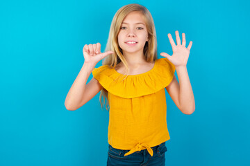 Caucasian kid girl wearing yellow T-shirt against blue wall showing and pointing up with fingers number six while smiling confident and happy.