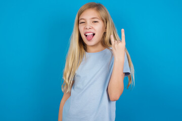 Portrait of a crazy beautiful Caucasian little girl wearing blue T-shirt over blue background showing tongue horns up gesture, expressing excitement of being on concert of band.