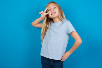 beautiful Caucasian little girl wearing blue T-shirt over blue background making v-sign near eyes. Leisure, coquettish, celebration, and flirt concept.