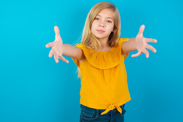 Caucasian kid girl wearing yellow T-shirt against blue wall looking at the camera smiling with open arms for hug. Cheerful expression embracing happiness.