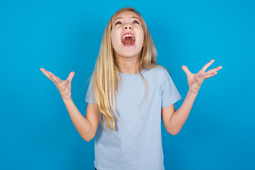 beautiful Caucasian little girl wearing blue T-shirt over blue background crying and screaming. Human emotions, facial expression concept. Screaming, hate, rage.
