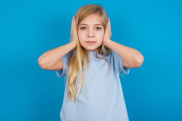 Fototapeta premium Frustrated beautiful Caucasian little girl wearing blue T-shirt over blue background plugging ears with hands does not wanting to listen hard rock, noise or loud music.