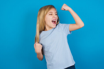 Fototapeta premium beautiful Caucasian little girl wearing blue T-shirt over blue background celebrating a victory punching the air with his fists and a beaming toothy smile