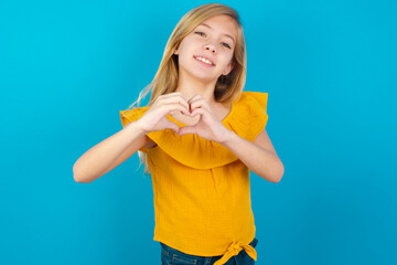 Caucasian kid girl wearing yellow T-shirt against blue wall smiling in love doing heart symbol shape with hands. Romantic concept.