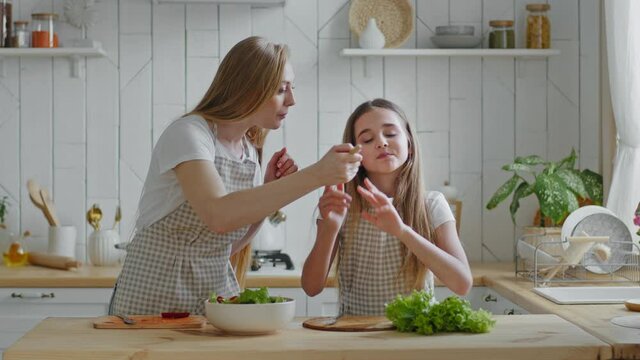Mother and daughter cook natural vegetable salad, mommy feeding baby from wooden spoon, girl eating delicious dish rejoices gives high five to woman jumping with happiness, family hugging in kitchen