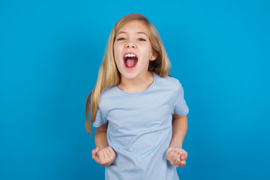 Joyful excited lucky beautiful Caucasian little girl wearing blue T-shirt over blue background cheering, celebrating success, screaming yes with clenched fists
