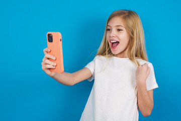 Happy cheerful beautiful Caucasian little girl wearing white T-shirt over blue background receiving good news via e-mail and celebrating success while standing and looking at mobile phone.