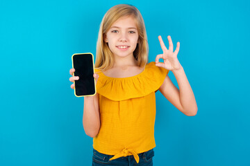 Caucasian kid girl wearing yellow T-shirt against blue wall holding in hands cell showing ok-sign
