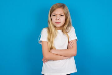 Gloomy dissatisfied beautiful Caucasian little girl wearing white T-shirt over blue background looks with miserable expression at camera from under forehead, makes unhappy grimace