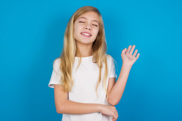 Overjoyed successful beautiful Caucasian little girl wearing white T-shirt over blue background raises palm and closes eyes in joy being entertained by friends