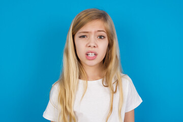 Portrait of dissatisfied beautiful Caucasian little girl wearing white T-shirt over blue background...