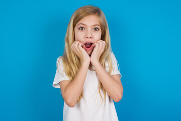 Speechless beautiful Caucasian little girl wearing white T-shirt over blue background keeps hands near opened mouth reacts to shocking news stares wondered at camera