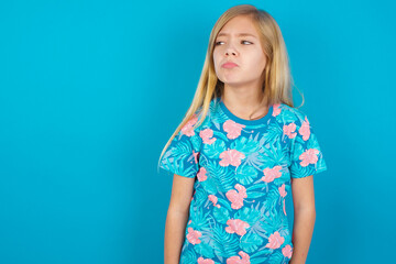 Dissatisfied Caucasian kid girl wearing hawaiian T-shirt against blue wall purses lips and has unhappy expression looks away stands offended. Depressed frustrated model.