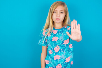 Caucasian kid girl wearing hawaiian T-shirt against blue wall shows stop sign prohibition symbol keeps palm forward to camera with strict expression