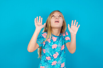 Caucasian kid girl wearing hawaiian T-shirt against blue wall keeps palms forward and looks with fright above on ceiling tries to defense herself from invisible danger opens mouth.